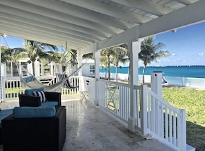 A covered outdoor seating area with wicker furniture and a hammock, overlooking a beach with palm trees and a clear blue sky. - Blue Beach Front Home at Resort World (North Bimini)