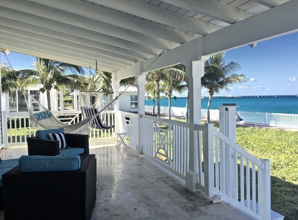 A covered outdoor seating area with wicker furniture and a hammock, overlooking a beach with palm trees and a clear blue sky.