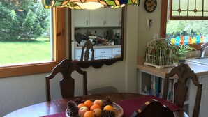 Un comedor con una lámpara colgante de vitral, una mesa de madera con una cesta de frutas y vista a la cocina por una ventana.