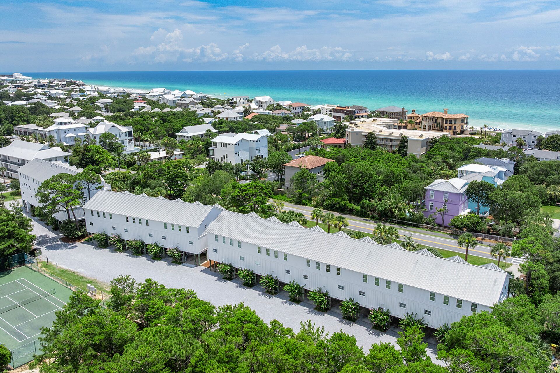 An aerial view of a coastal residential area with houses, a tennis court, and a view of the ocean.