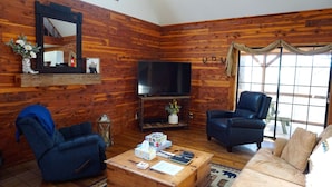 Una sala de estar acogedora con paredes de madera, televisor, dos sillones, un sofá y una mesa de centro con diversos objetos.