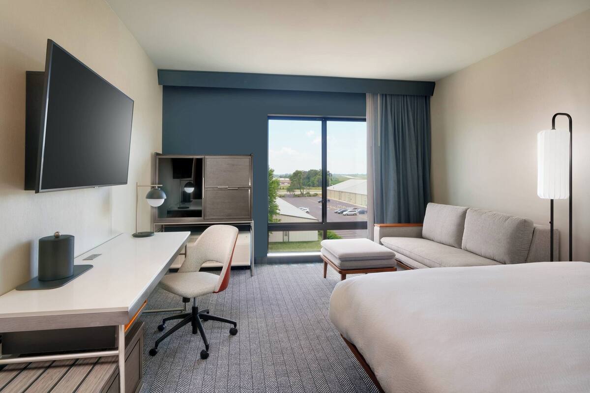 A hotel room with a bed, a desk with a chair, a TV, a large window with a view of the outside, and a lamp.