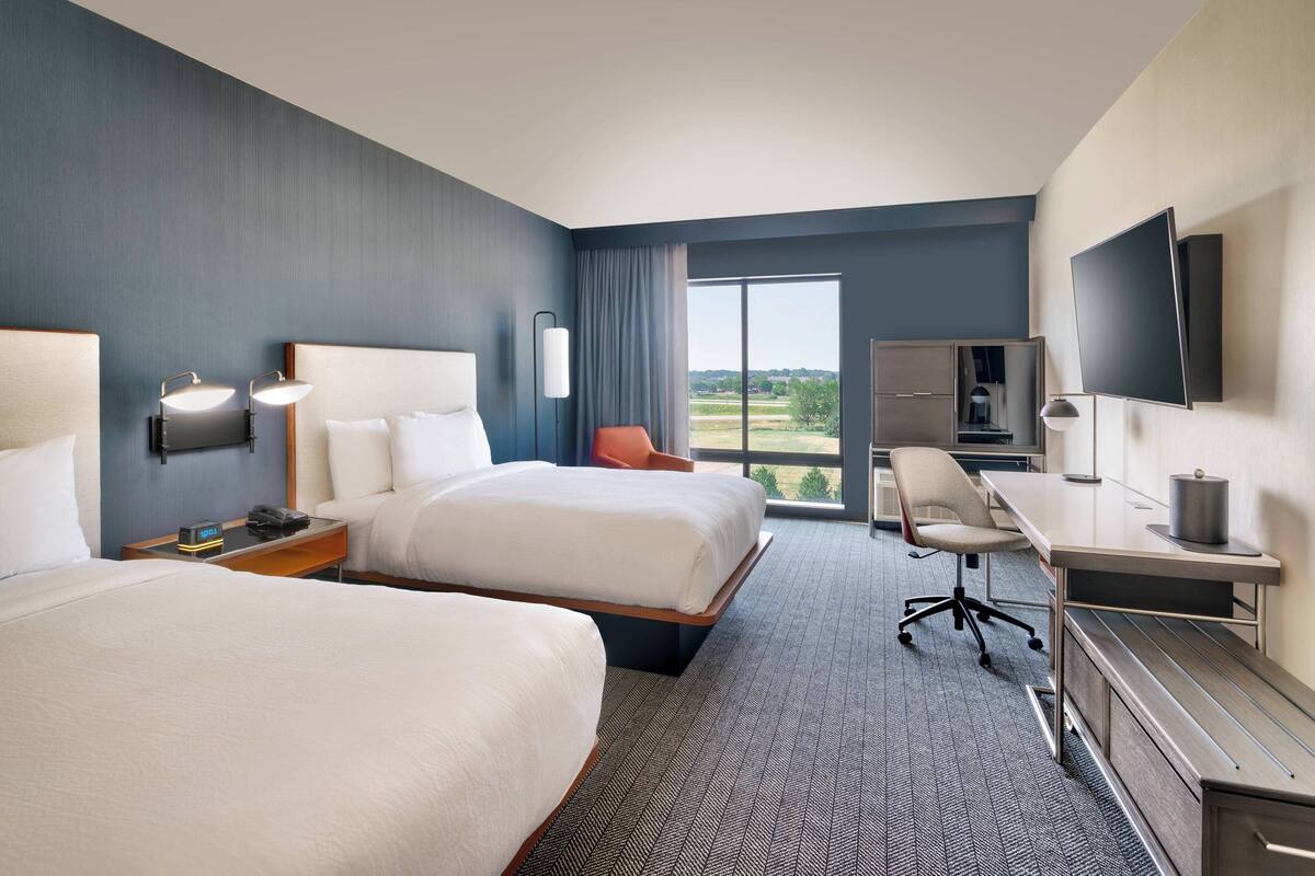 A hotel room with two beds, a desk, a chair, a TV, and a large window with a view.