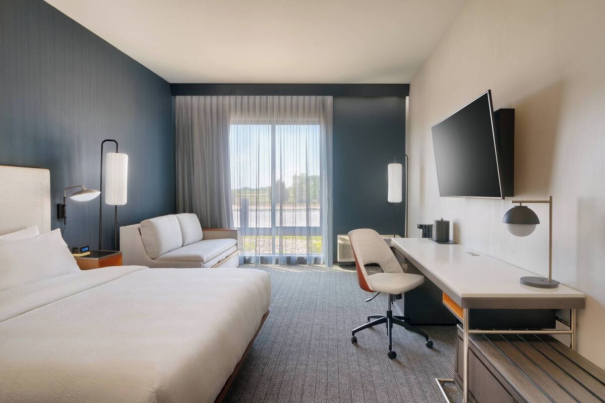 A hotel room with a large bed, a desk, a chair, a TV, and a window with curtains.