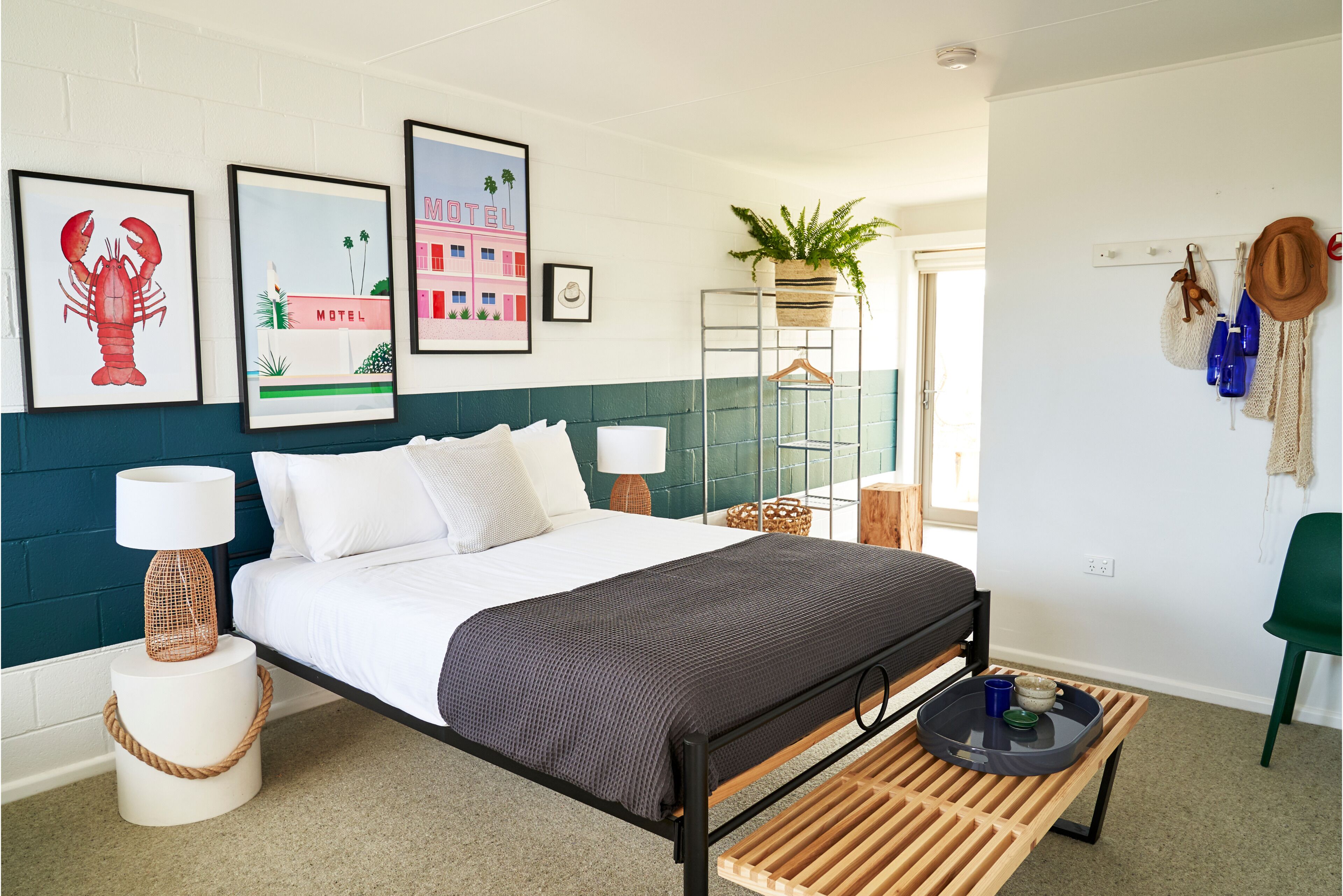 A neatly arranged bedroom with a bed, bedside lamps, a tray table, and wall art depicting a lobster and a motel.