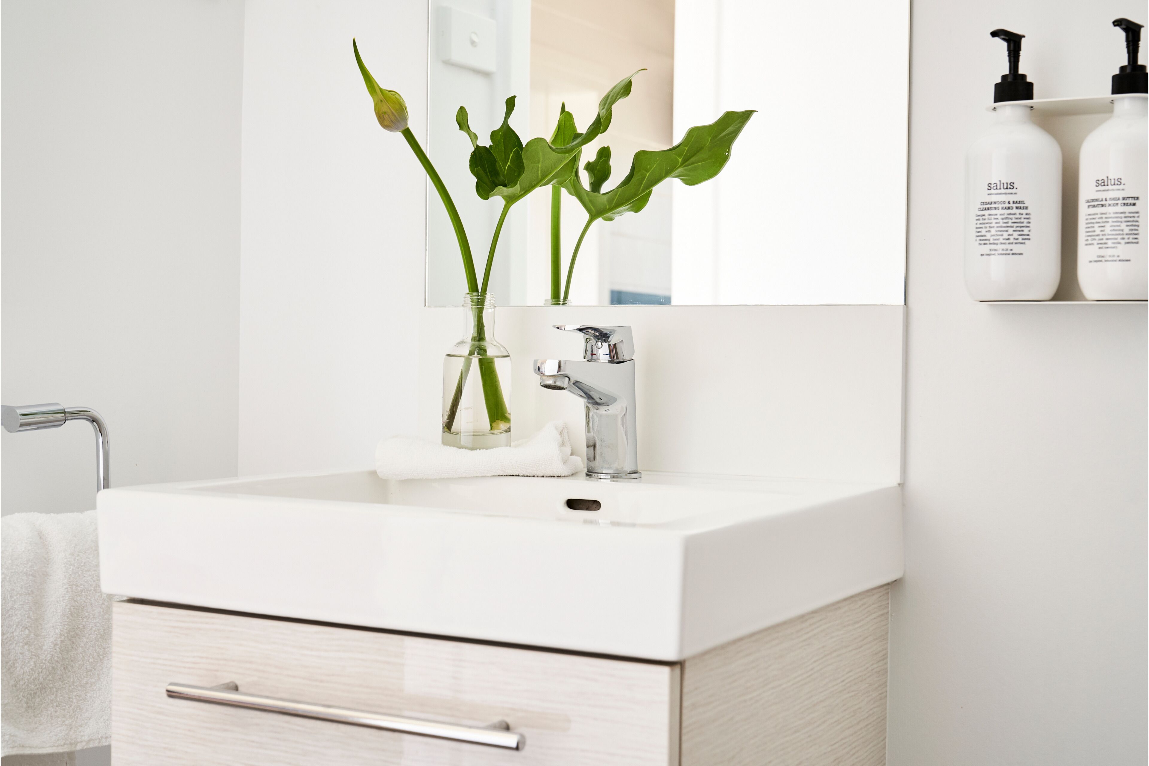Ein modernes Badezimmer mit einem weißen Waschbecken, einer Glasvase mit grünen Blättern und einem Spiegel über dem Waschbecken.
