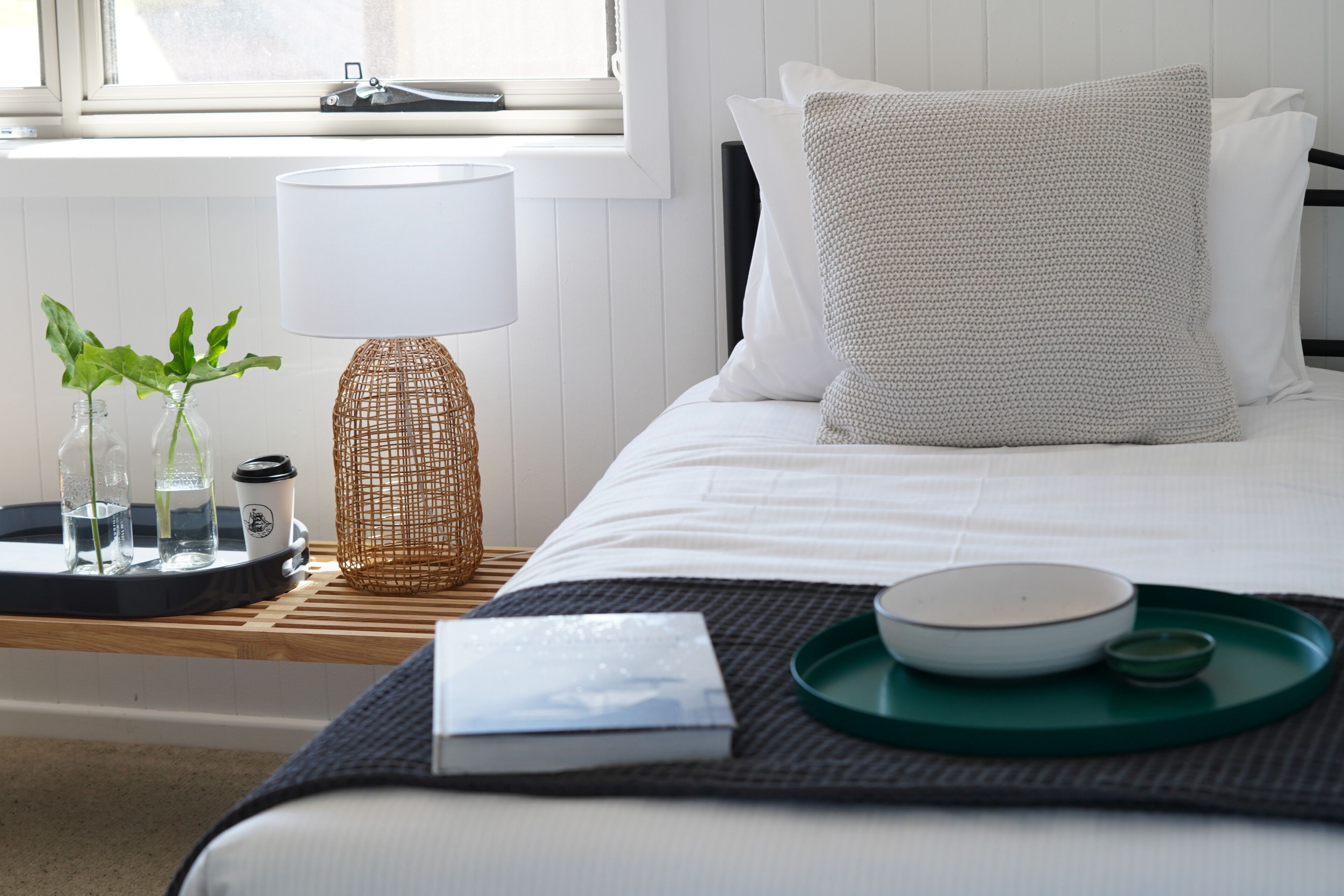 A neatly made bed with a white pillow and a textured pillow, a bedside table with a lamp, a tray with a bowl and a cup, and a small plant in a glass jar.