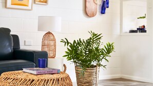 Un salón con un sofá negro, un reposapiés de mimbre con un libro y una taza azul, una lámpara trenzada y una planta en maceta.