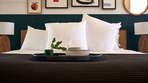 Una cama bien tendida con almohadas blancas, una bandeja con una taza verde y un plato, y una obra de arte en la pared.
