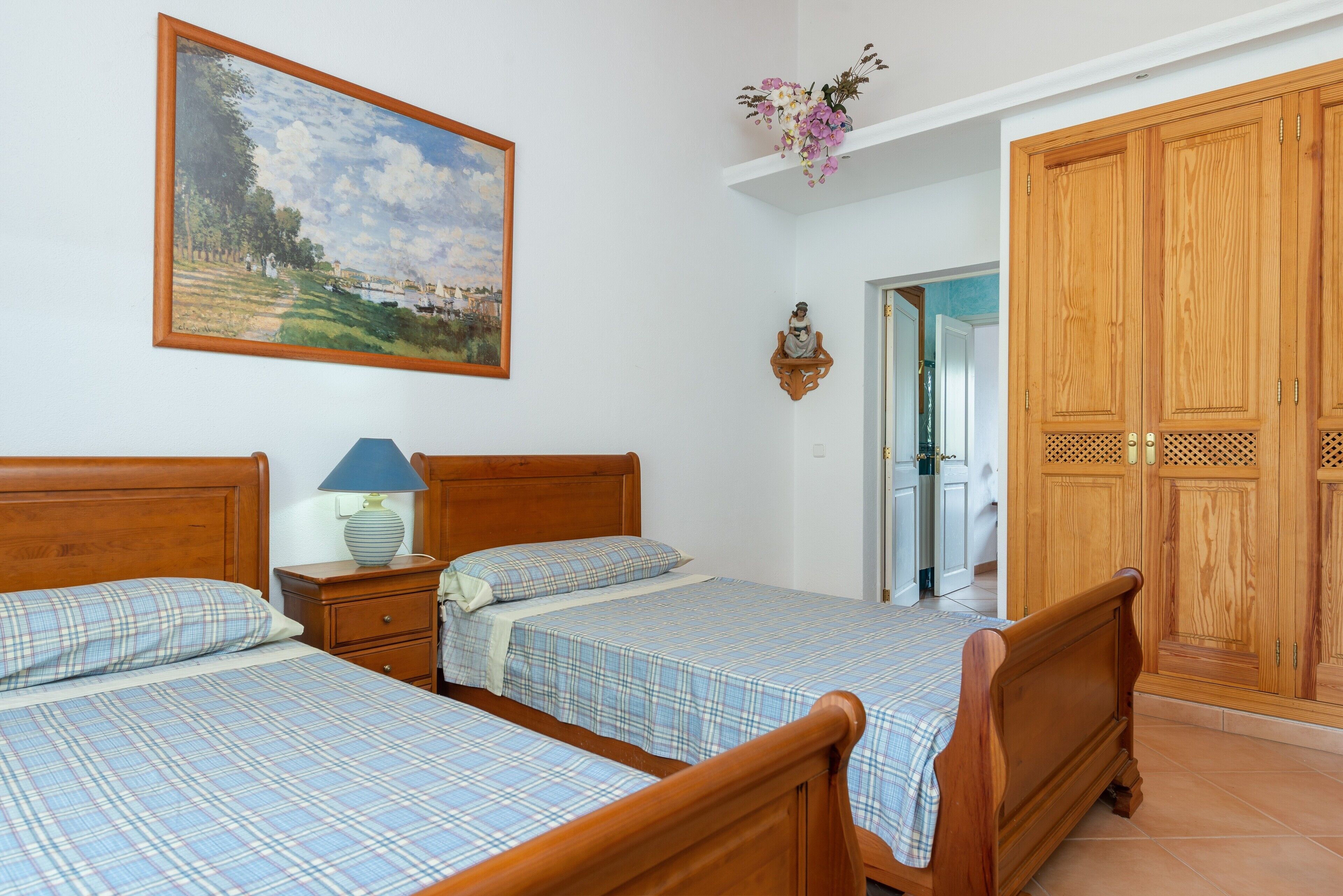A bedroom with two wooden beds, a nightstand with a lamp, a painting on the wall, and a wooden wardrobe.