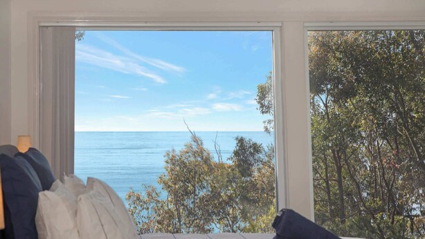 Ein Schlafzimmer mit großem Fenster mit Blick auf ein Gewässer und Bäume. Auf dem Bett liegen Kissen und ein zusammenrolltes Handtuch.