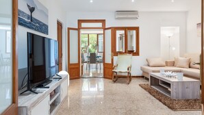 A modern living room with a flat-screen TV, a wooden entertainment unit, a beige sofa, a coffee table, and a dining area visible through an open door.