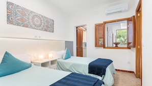 A bedroom with two beds, a nightstand, a wall-mounted air conditioner, and a window with a view of greenery.