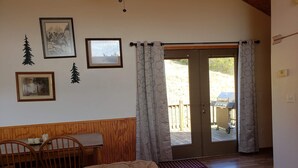 Une pièce comprenant un lit, un banc en bois et une table avec des bougies. Il y a deux fenêtres avec des rideaux, et une porte qui donne sur l’extérieur.