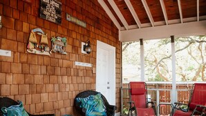 A covered porch with wooden walls, a white door, and a view of trees. There are wicker chairs and a red chair.