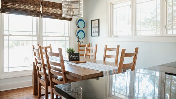 A dining area with a wooden table, chairs, a hanging light fixture, and a view through large windows.