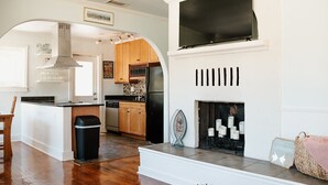 A spacious living room with a fireplace, a kitchen with wooden cabinets, and a large TV mounted above the fireplace.