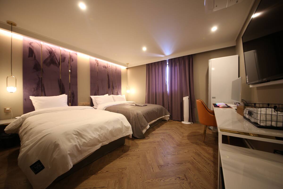 Habitación de hotel con dos camas, un escritorio con silla, un televisor y un ventanal con cortinas.
