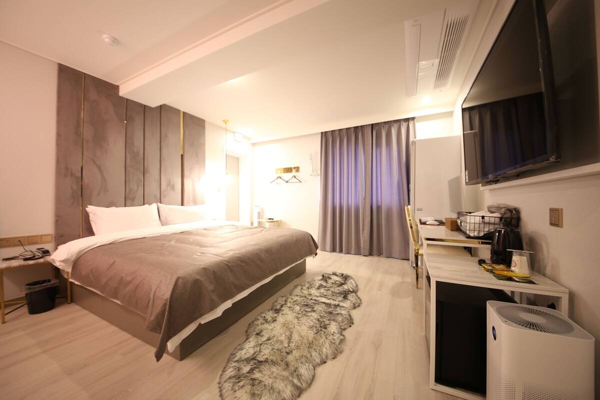 Una habitación de hotel moderna con una cama grande, un televisor de pantalla plana, un escritorio con una silla y un ventanal con cortinas.