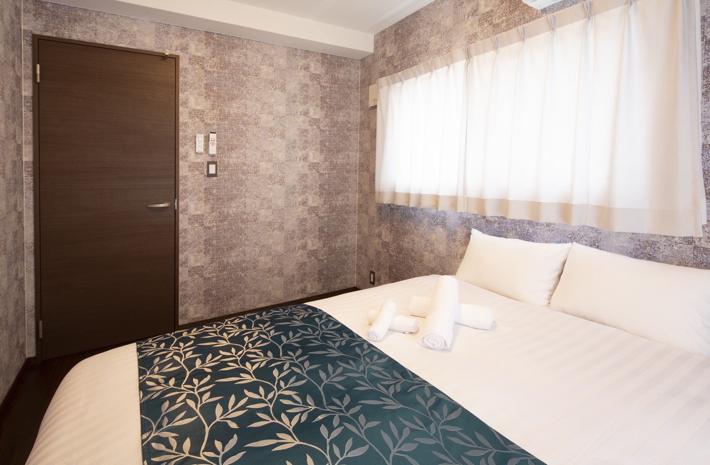 A hotel room with a large bed, a dark wooden door, textured walls, and a window with sheer curtains.
