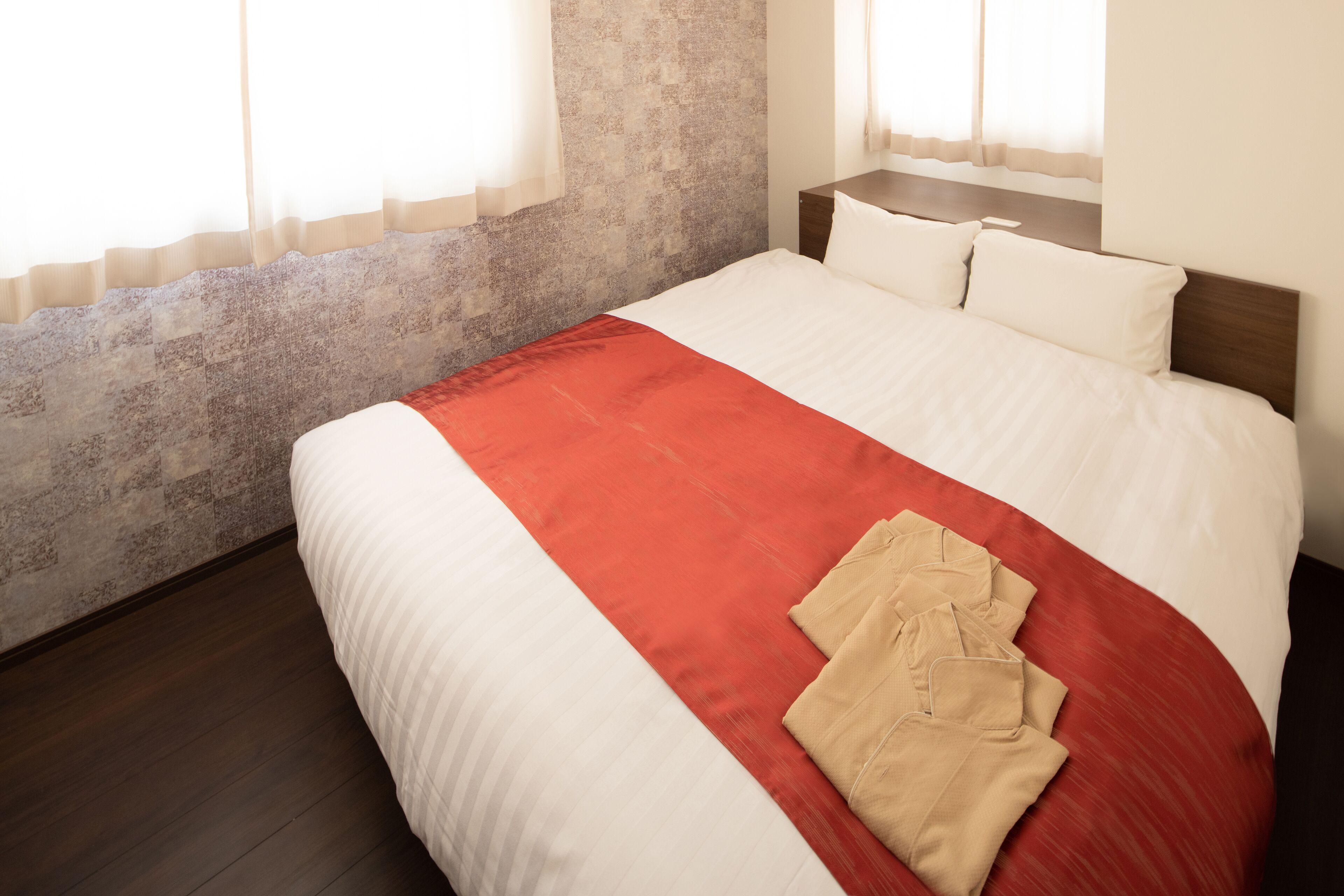 Una cama bien tendida con una colcha roja y blanca, un cabecero de madera y un par de pantalones beige doblados sobre la cama.