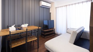 Habitación de hotel con una mesa de comedor de madera, sillas, televisor de pantalla plana, sofá y un amplio ventanal con cortinas.