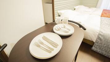 Habitación de hotel ordenada con una mesa de comedor preparada para dos personas, una cama con ropa blanca y una mesita con una taza y un platillo.
