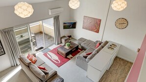 A modern living room with a sofa set, a coffee table, and a kitchen area with a sink. There are large windows and a view of the outside. - Razorback 15 - Central Jindabyne CBD Unit (Jindabyne)