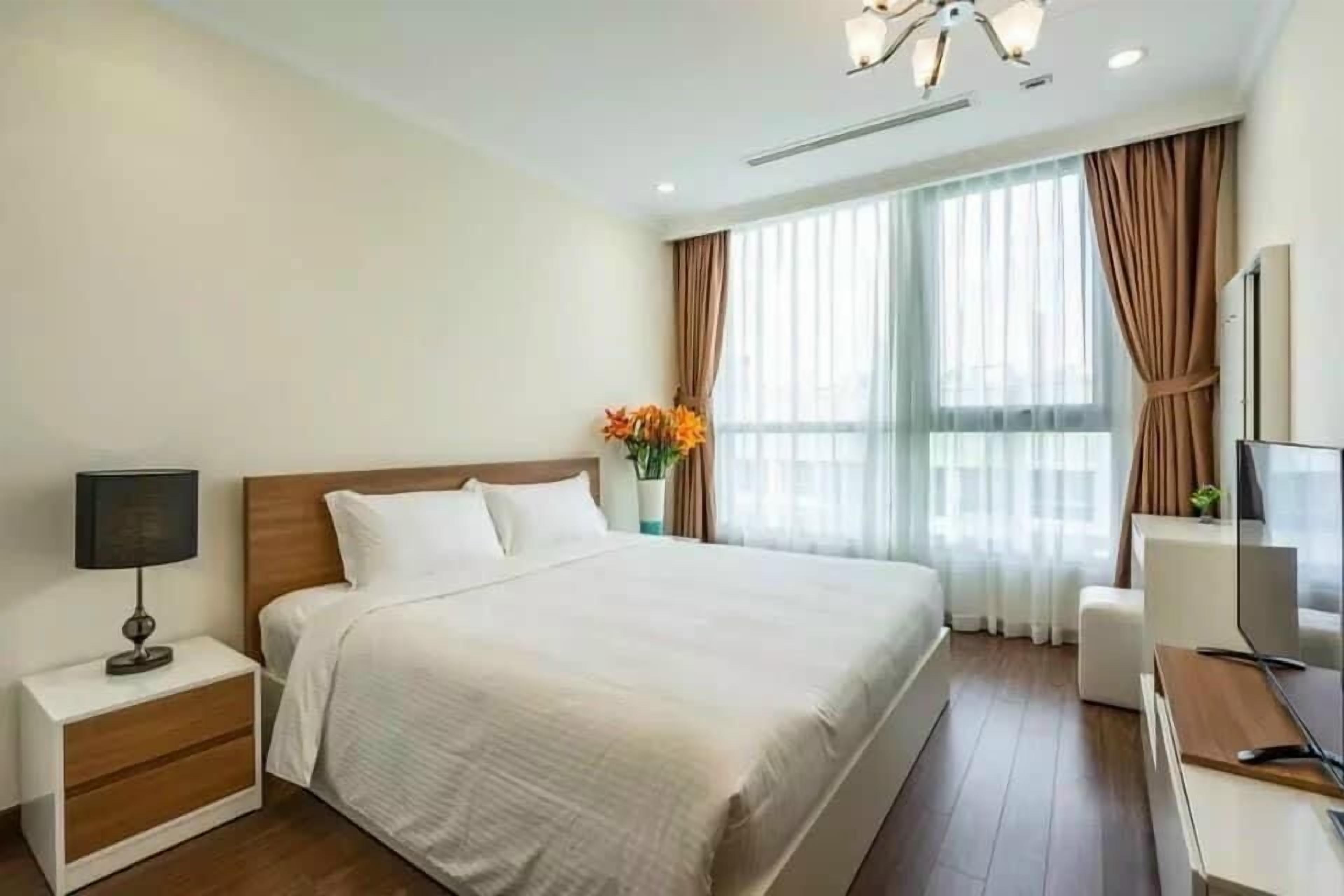 Una habitación de hotel con una cama grande, una mesita de noche con lámpara, un televisor y una ventana con cortinas.