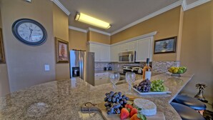 A kitchen with a granite countertop, a clock, white cabinets, a refrigerator, a microwave, and a fruit bowl. - Penthouse@Shores Of Panama 🏖Million Dollar View! (Panama City Beach)