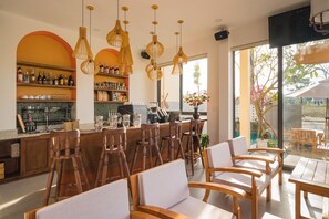 Un área de bar moderna con sillas y mesas de madera, una barra con estantes llenos de botellas y un amplio ventanal con vistas al exterior.