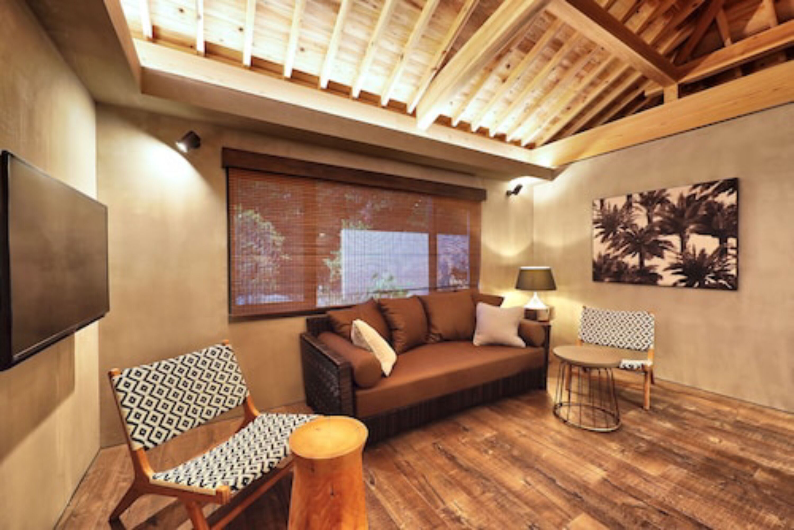 A cozy living area with a sofa, patterned chair, and round table, featuring a wooden floor and a large window with blinds.