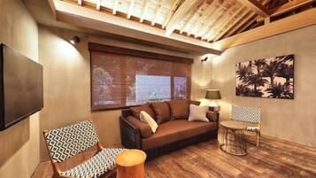 A cozy living area with a sofa, patterned chair, and round table, featuring a wooden floor and a large window with blinds.
