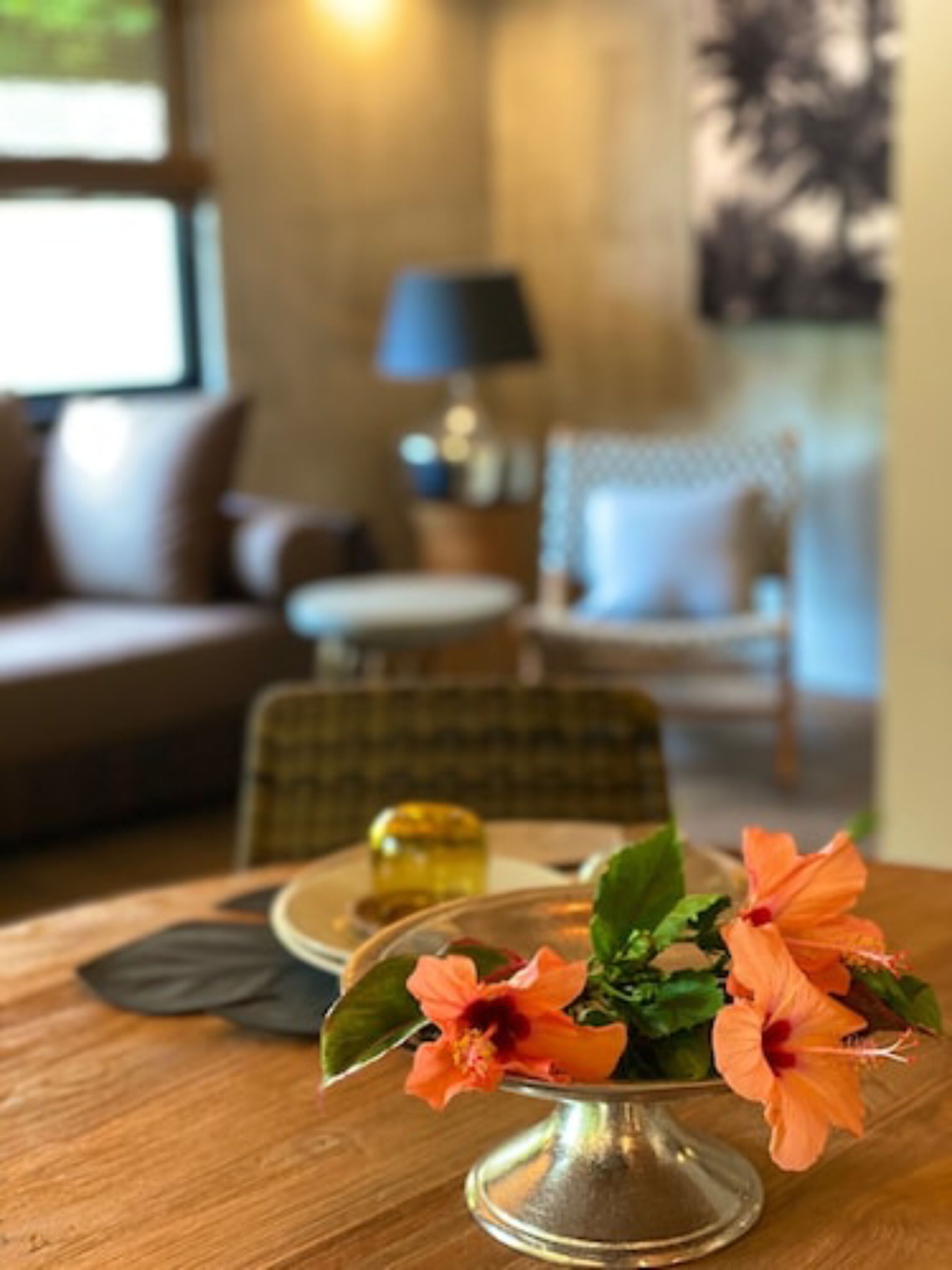 Una mesa de comedor con un centro de mesa floral, una lámpara y una silla en una habitación acogedora con un sofá y un cuadro en la pared.