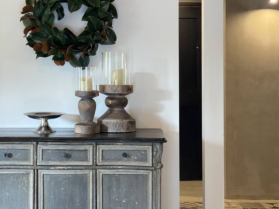 A vintage-style dresser with two glass candle holders and a decorative wreath on the wall behind it.