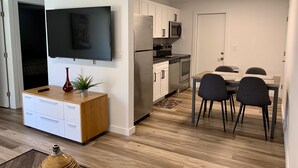 Cocina moderna con armarios blancos, un refrigerador de acero inoxidable y un microondas. Hay un comedor con una mesa de madera y cuatro sillas. Un televisor de pantalla plana está montado en la pared sobre la encimera de la cocina.