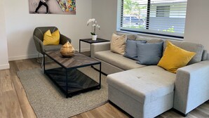 Una sala de estar moderna con un sofá gris, una mesa de centro negra y un sillón gris con cojines amarillos. Hay un cuadro en la pared y una ventana con persianas.