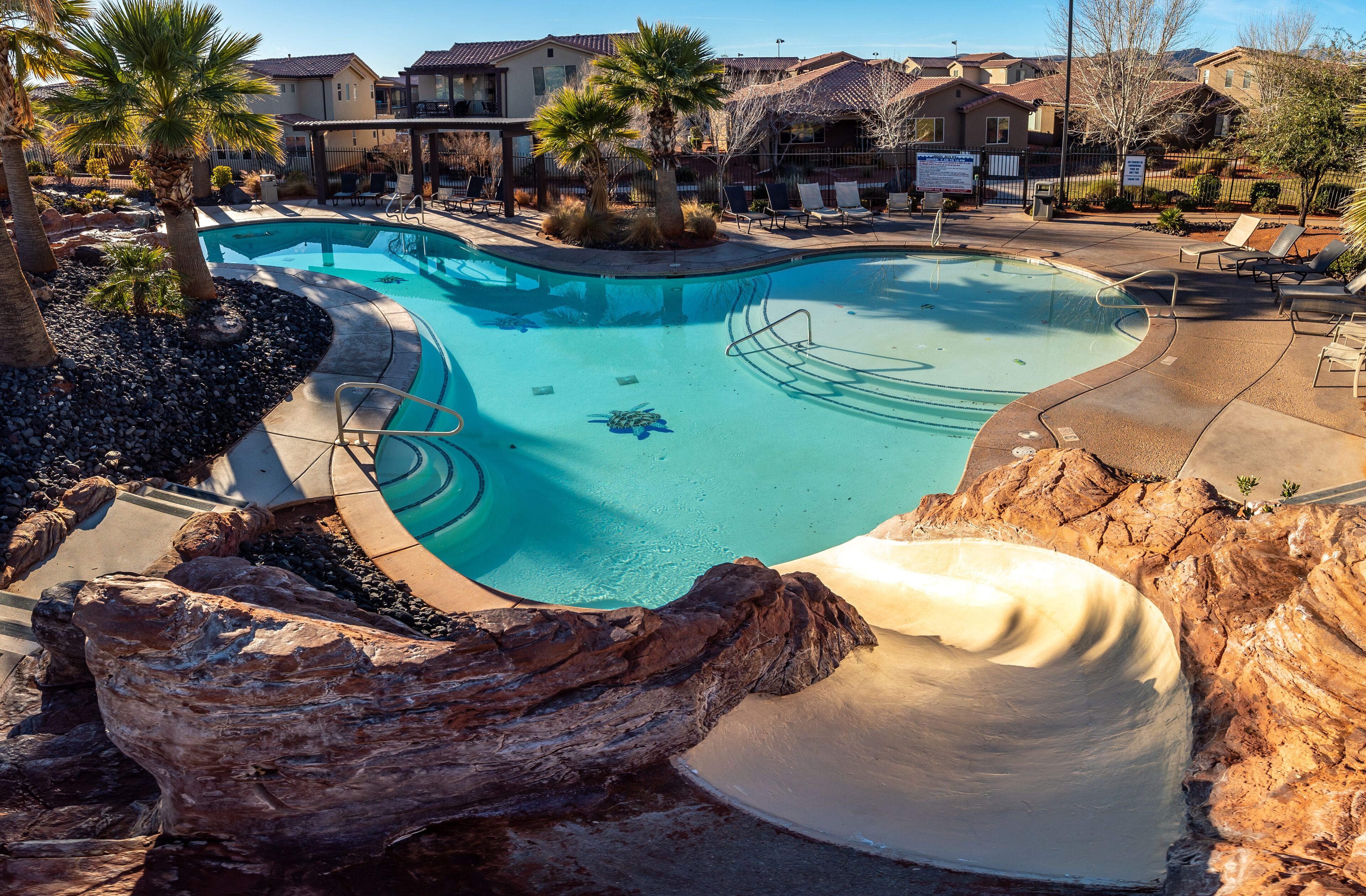 A swimming pool with a slide, surrounded by lounge chairs and palm trees.