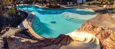 A swimming pool with a slide, surrounded by lounge chairs and palm trees.