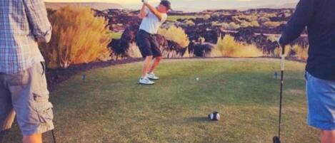 Ein Golfplatz mit einem Spieler im Schwung, einer weiteren Person, die zusieht, und einer dritten Person, die einen Golfschläger hält.