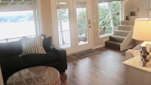 A living room with a dark-colored sofa, a round woven ottoman, and a lamp on a table. There is a set of stairs leading to a balcony with a railing. - Hamilton Happy Place (million dollar view) - Lake Retreat (Hot Springs)