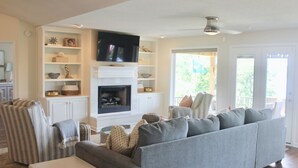 A living room with a gray sofa, striped armchair, and a fireplace. Shelves with decorative items and a flat-screen TV are visible. - Hamilton Happy Place (million dollar view) - Lake Retreat (Hot Springs)