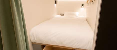 Une pièce de taille modeste, comprenant un lit, une petite table de chevet et un luminaire fixé au mur.