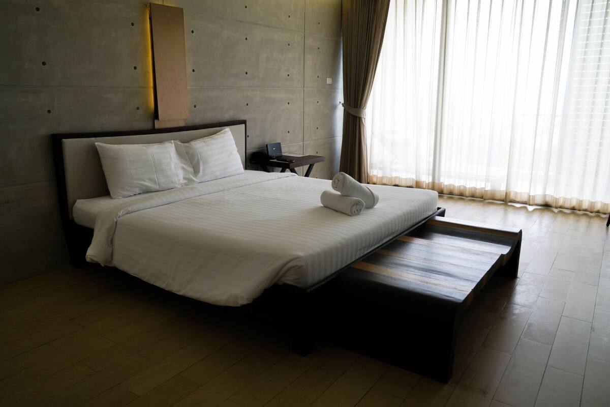 A modern hotel room with a large bed, a wooden bench, a desk, and a laptop.