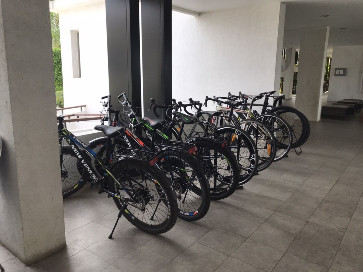 A row of bicycles parked indoors with a tiled floor and a view of a building outside.