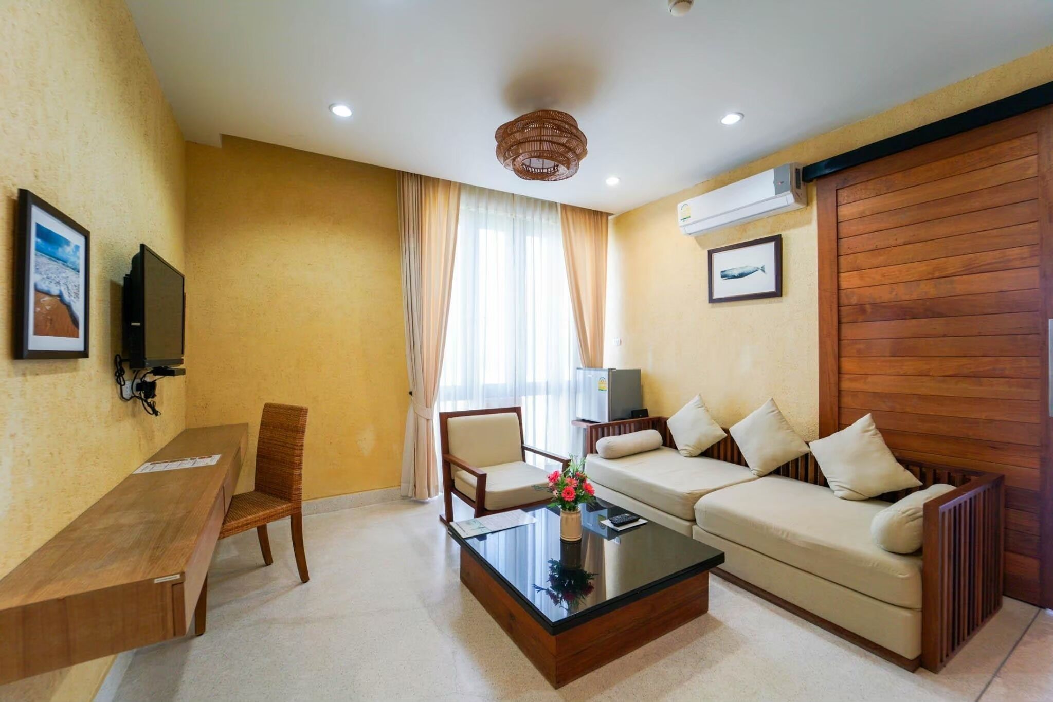 Habitación de hotel con sofá, una silla, una mesa de centro, televisor y ventana con cortinas.