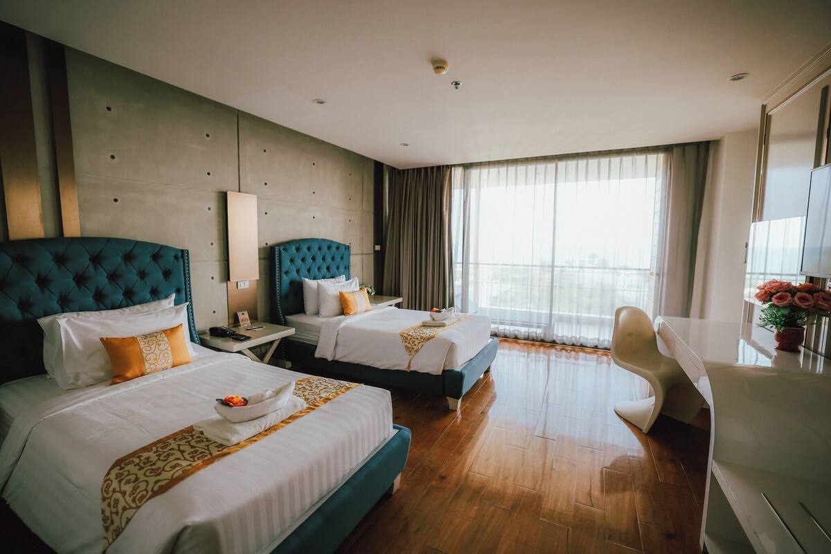 A hotel room with two beds, a desk, a chair, a TV, and a large window.