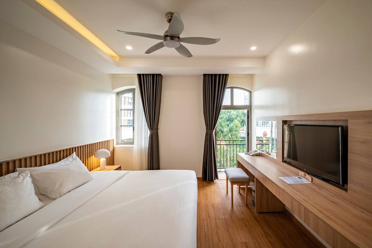 Une chambre d’hôtel avec un lit, un bureau avec une télévision, un ventilateur de plafond et une fenêtre avec des rideaux.