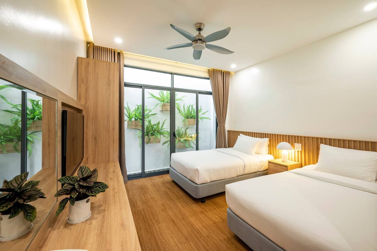 Une chambre d’hôtel avec deux lits, un ventilateur de plafond, une grande fenêtre avec des plantes et une table de chevet en bois.