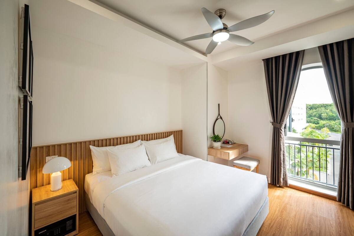 Une chambre d’hôtel avec un lit, une table de chevet avec une lampe, un ventilateur de plafond, une fenêtre avec des rideaux et une vue sur la verdure à l’extérieur.